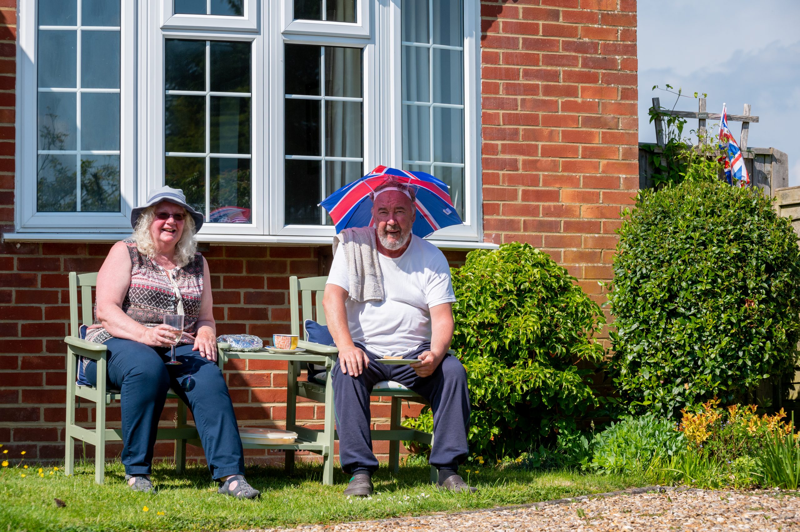 Two seniors sitting outside a brick house on a sunny day, wearing hats for sun protection, surrounded by greenery and flowers.