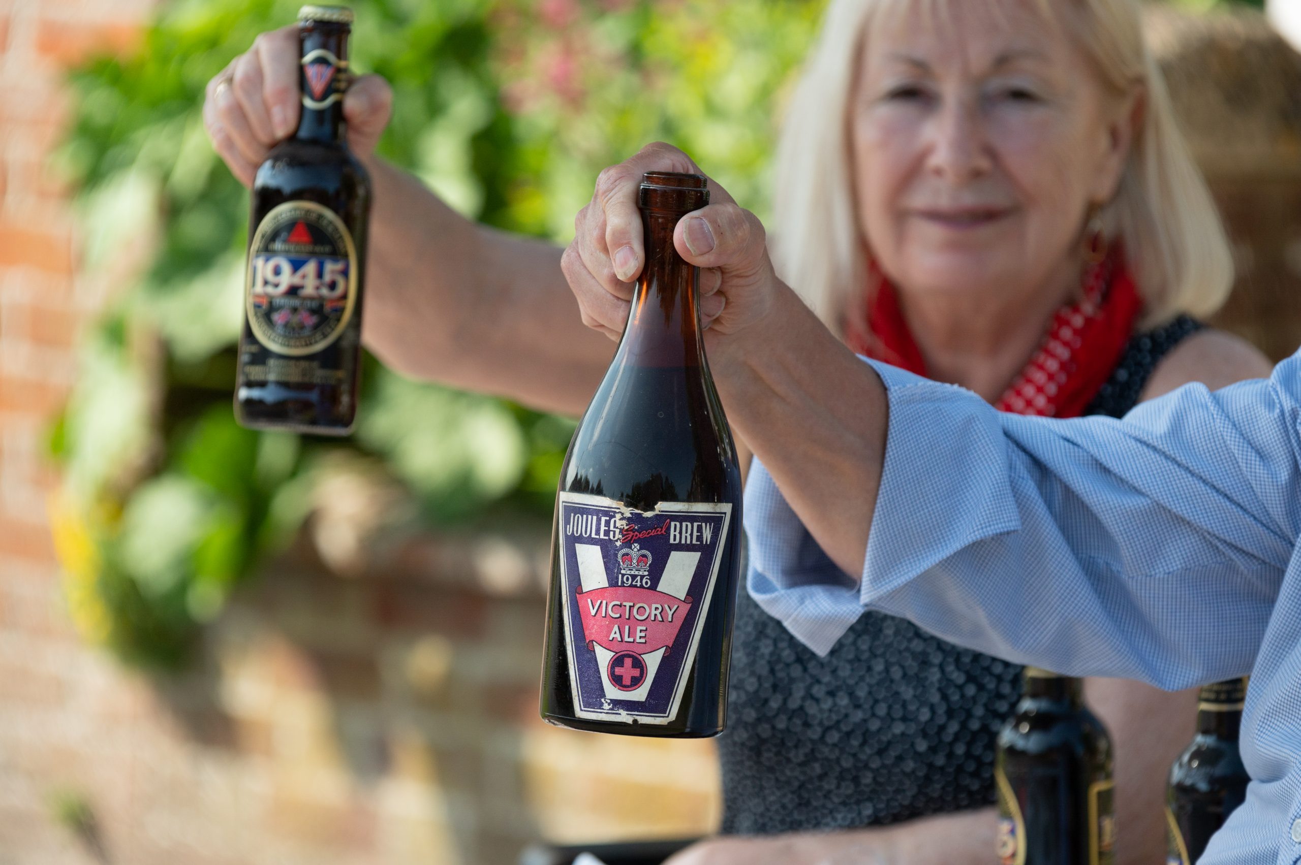 Two people holding craft beer bottles outdoors, showcasing a variety of labels. The setting features greenery, suggesting a casual and social atmosphere for beer tasting or brewing events.