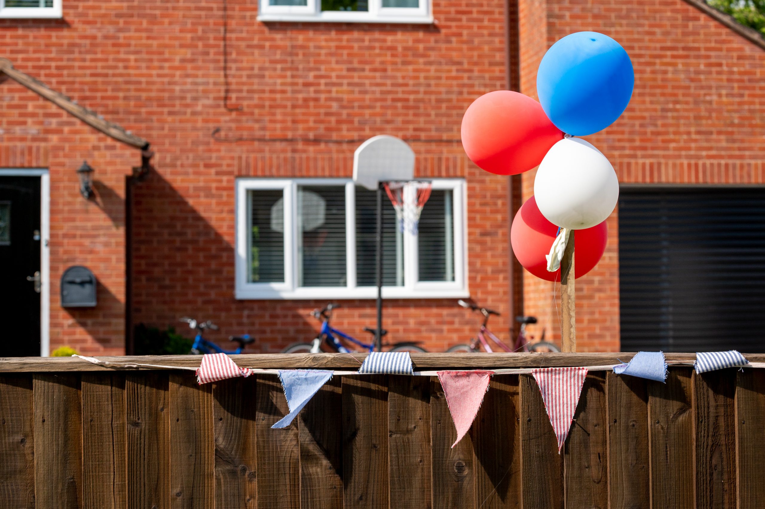 Colorful balloons in red, white, and blue float above a wooden fence, with festive bunting displayed nearby, set against the backdrop of a brick house featuring a basketball hoop and large windows.