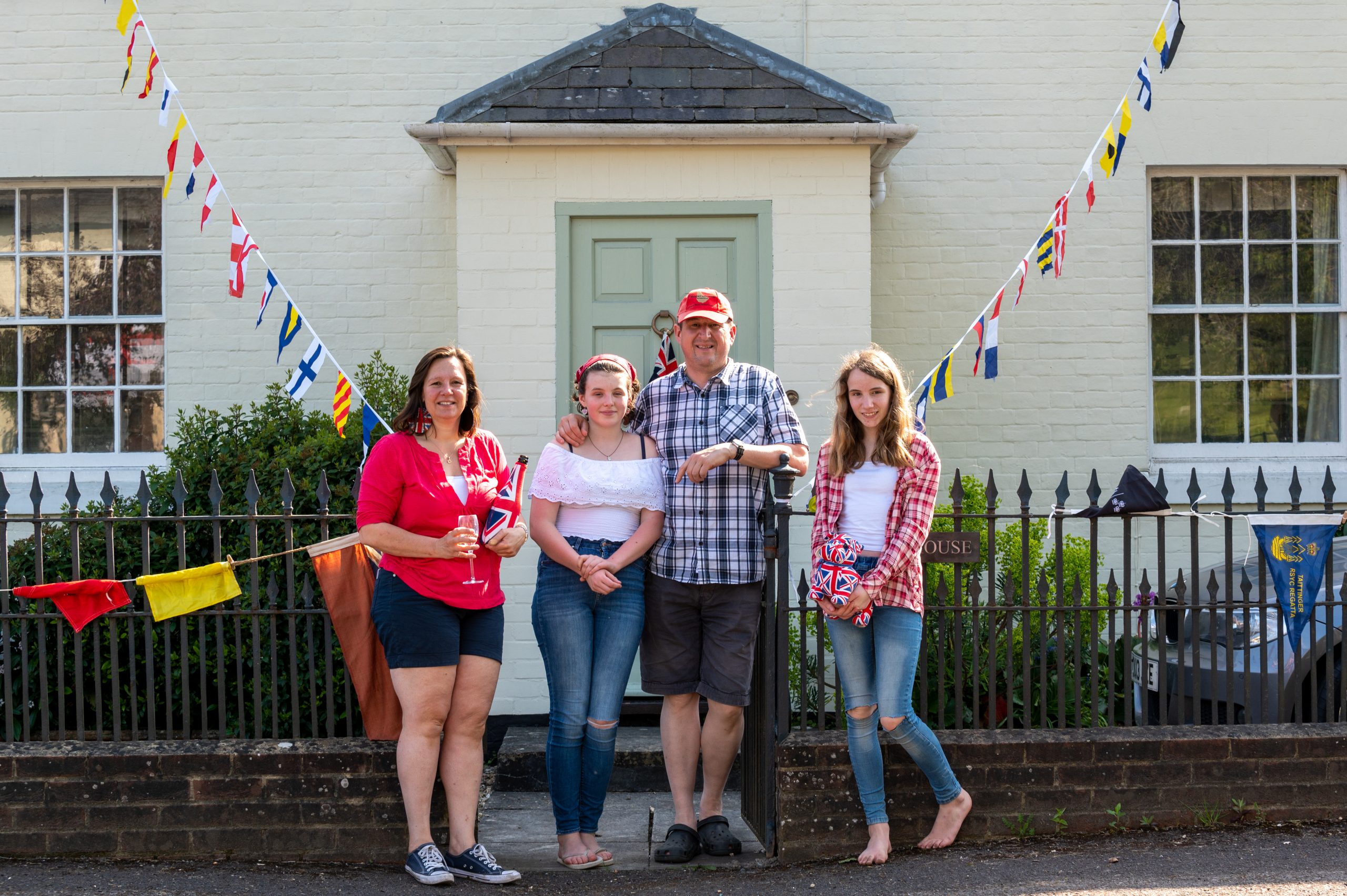 Group of four friends standing in front of a house decorated with colorful bunting, smiling and enjoying a sunny day. The scene captures a festive atmosphere, perfect for community events or gatherings.