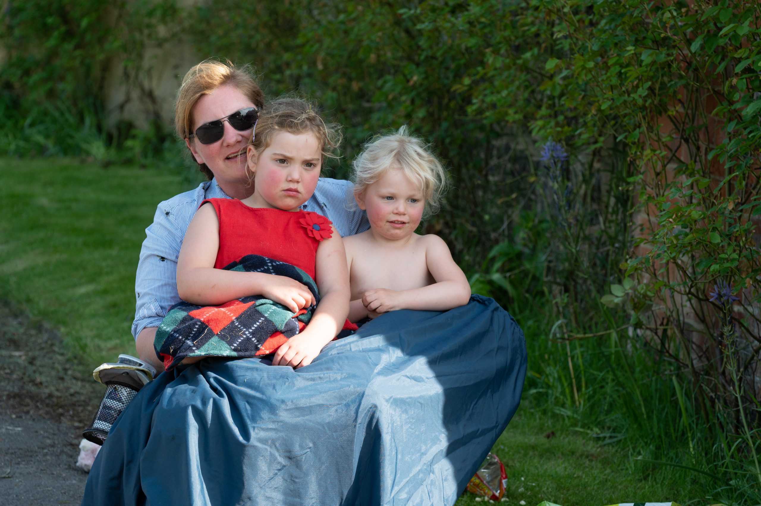 Woman seated outdoors with two children, one wearing a red shirt and the other shirtless, enjoying a sunny day on a path surrounded by greenery.