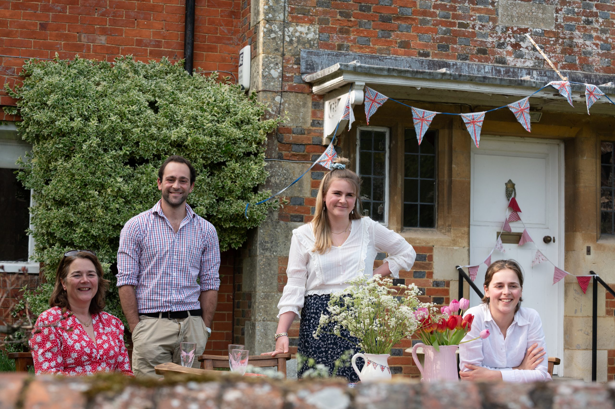 A group of four people smiling outdoors in front of a house, with a table featuring a floral arrangement. The setting is decorated with bunting, suggesting a festive atmosphere.