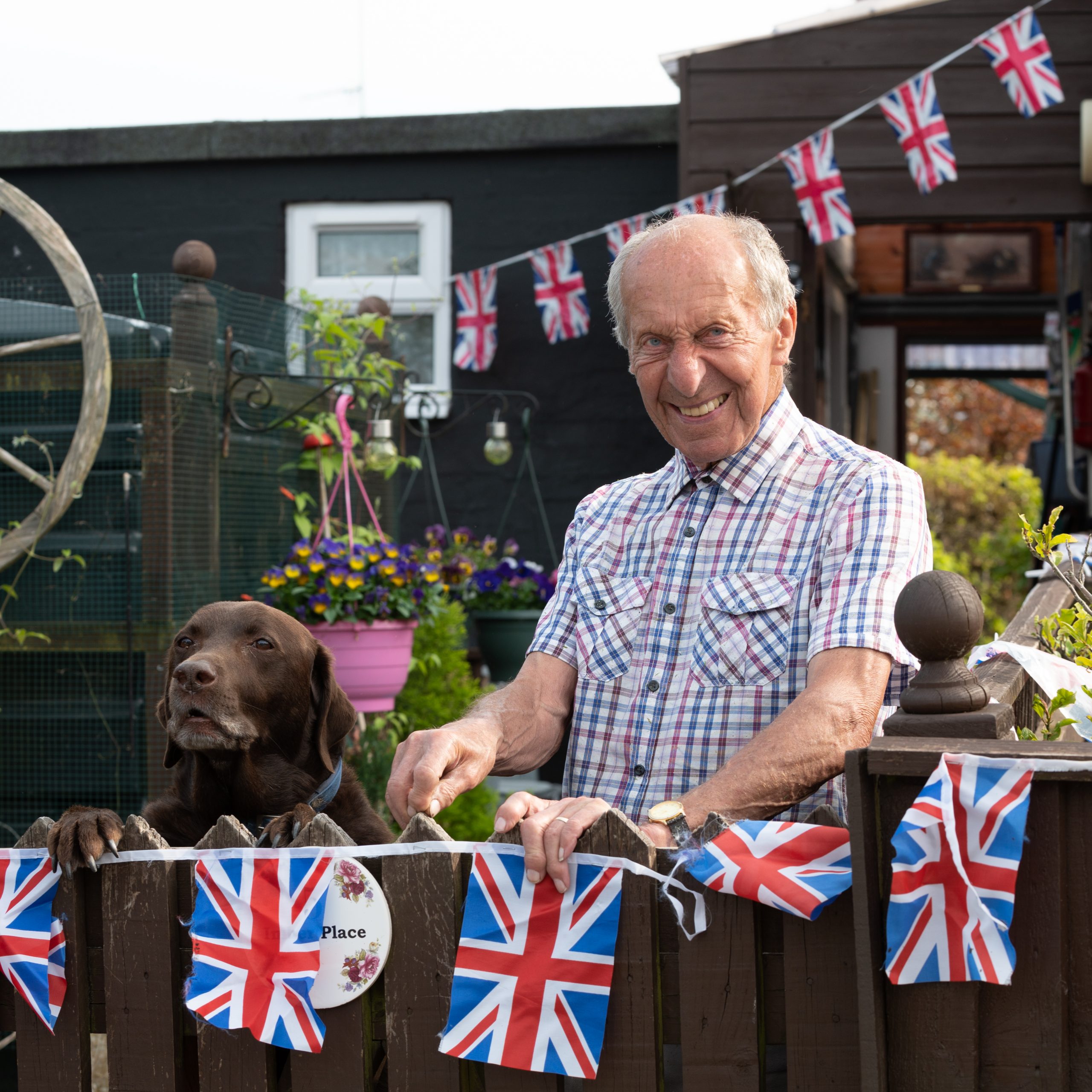 Elderly man standing by a wooden fence decorated with Union Jack flags, smiling alongside a brown dog in a garden setting.
