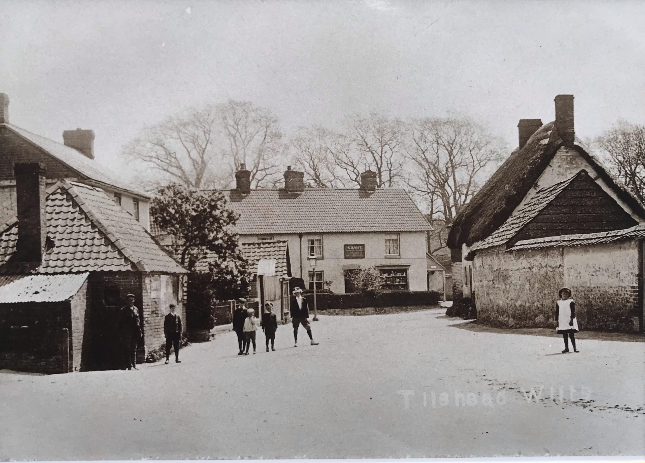 A historic black-and-white photograph depicting a village square with several thatched-roof houses. Children are seen playing in the foreground, and leafless trees are visible in the background.