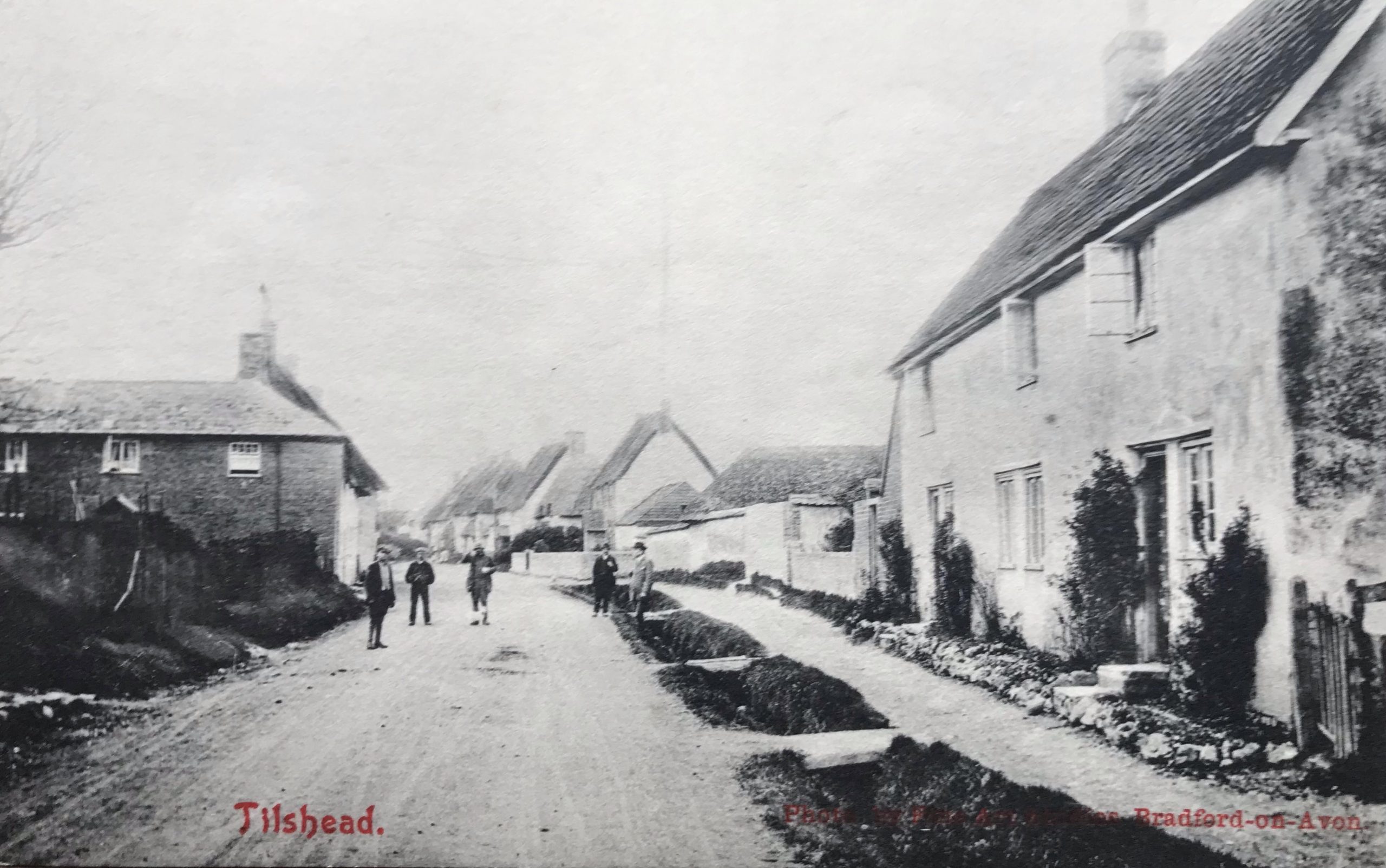 Historic black and white photograph of a rural street in a village, featuring traditional houses and people walking along the road, with a bridge visible in the background.