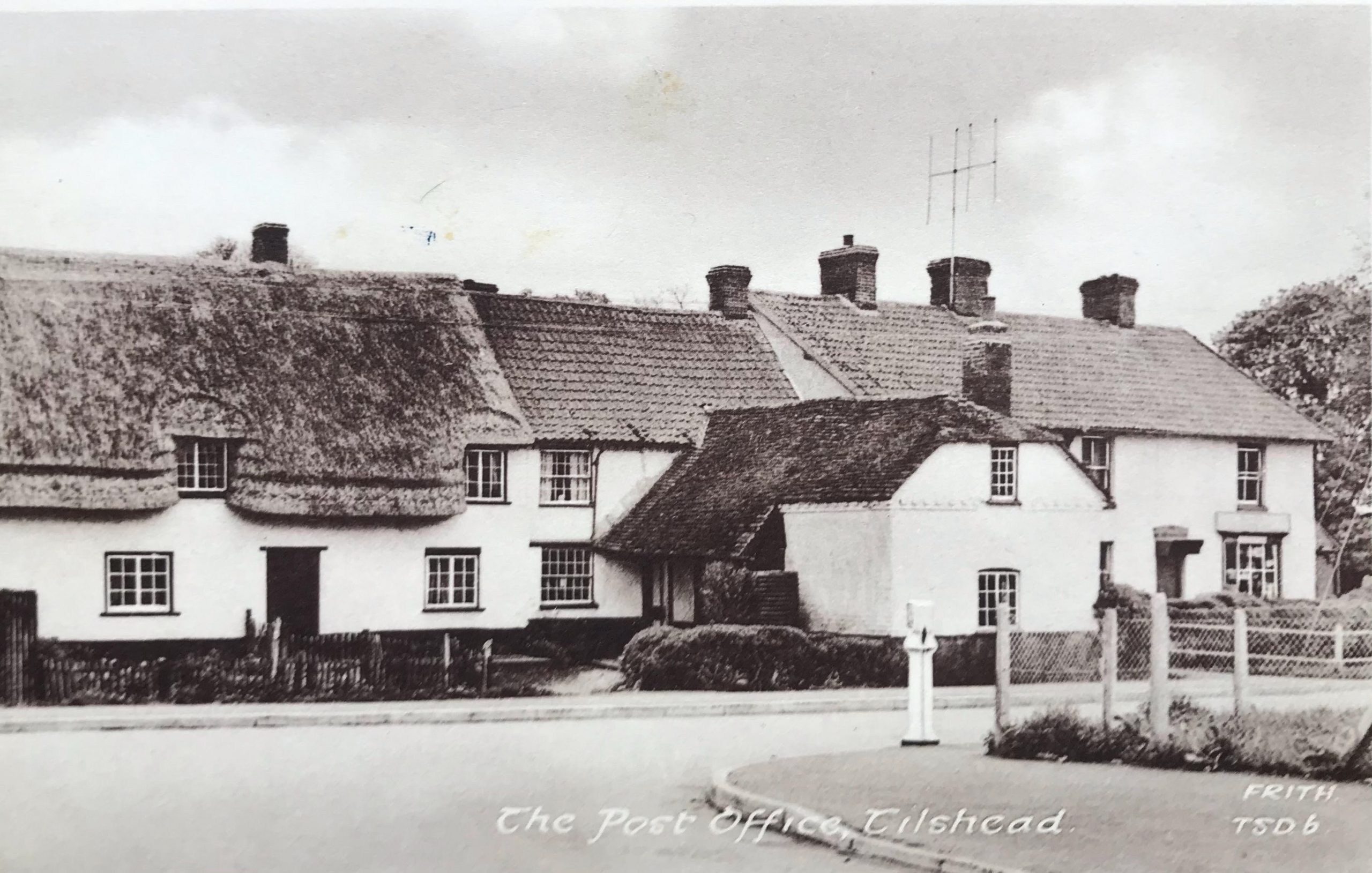 A historic black and white photograph of thatched-roof cottages lining a road, featuring a post office in Tilshead. White picket fences and a sign can be seen, set against a rural landscape.