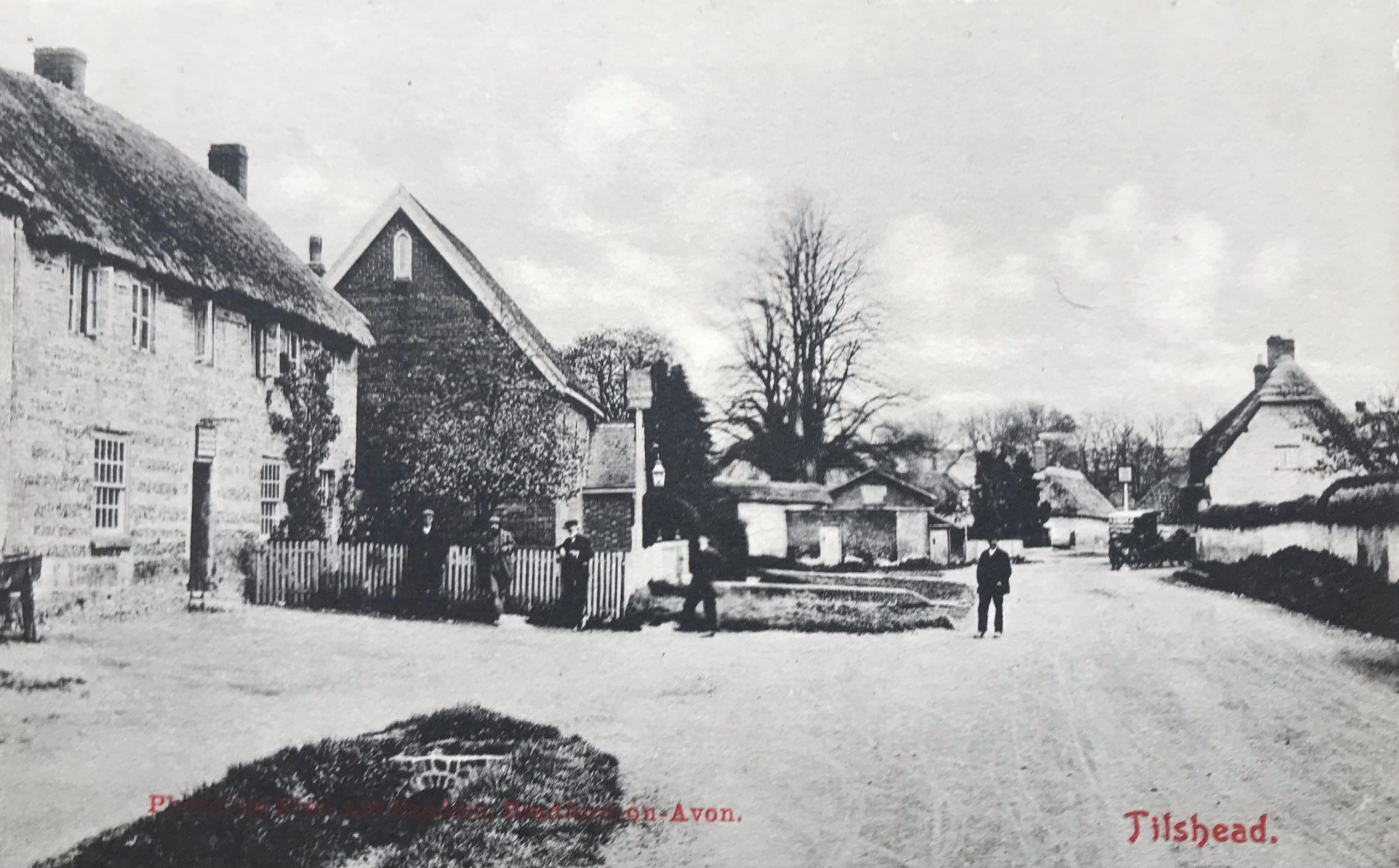 Black and white historical photograph of a quiet street in a small village, featuring traditional architecture, trees, and a person walking along the road, capturing the essence of rural life.