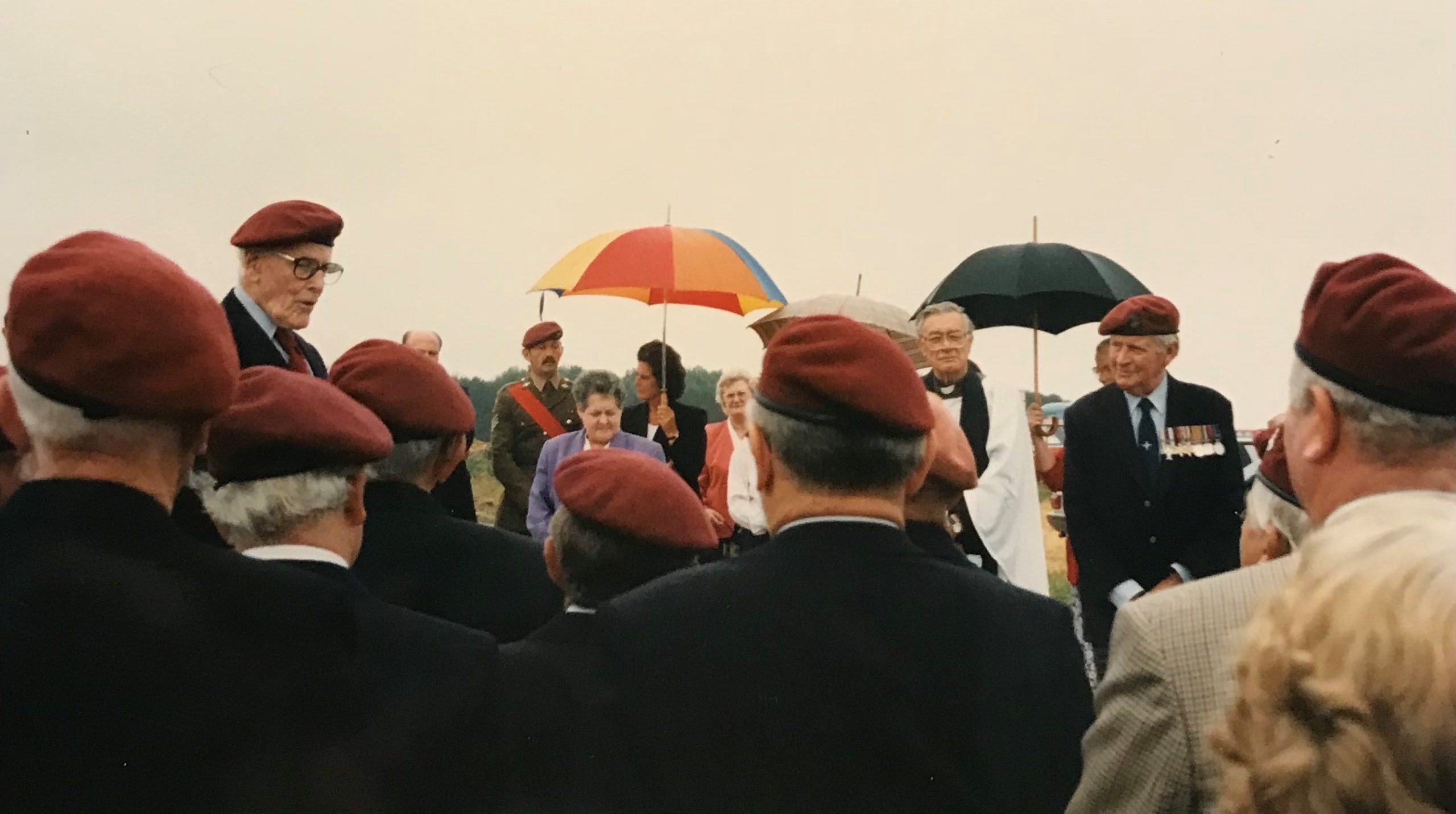 A gathering of people wearing red berets, some holding umbrellas, while listening to speakers at an outdoor event. The setting appears formal, likely commemorative.