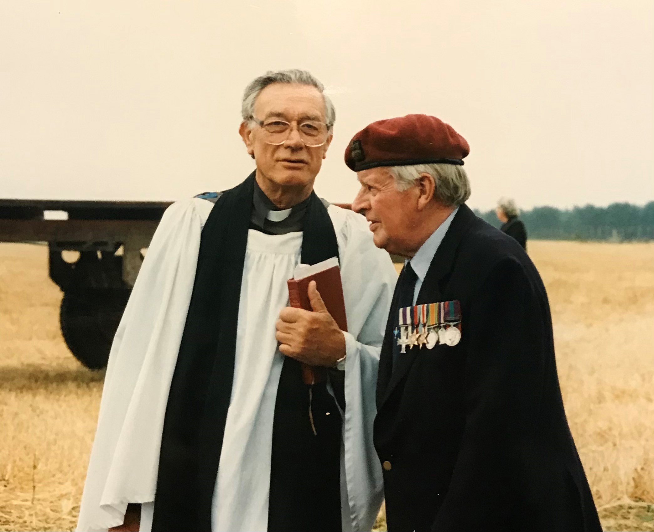 Two men engaged in conversation outdoors, one wearing a clerical robe and the other in military attire with medals and a beret, set against a backdrop of a field and military equipment.
