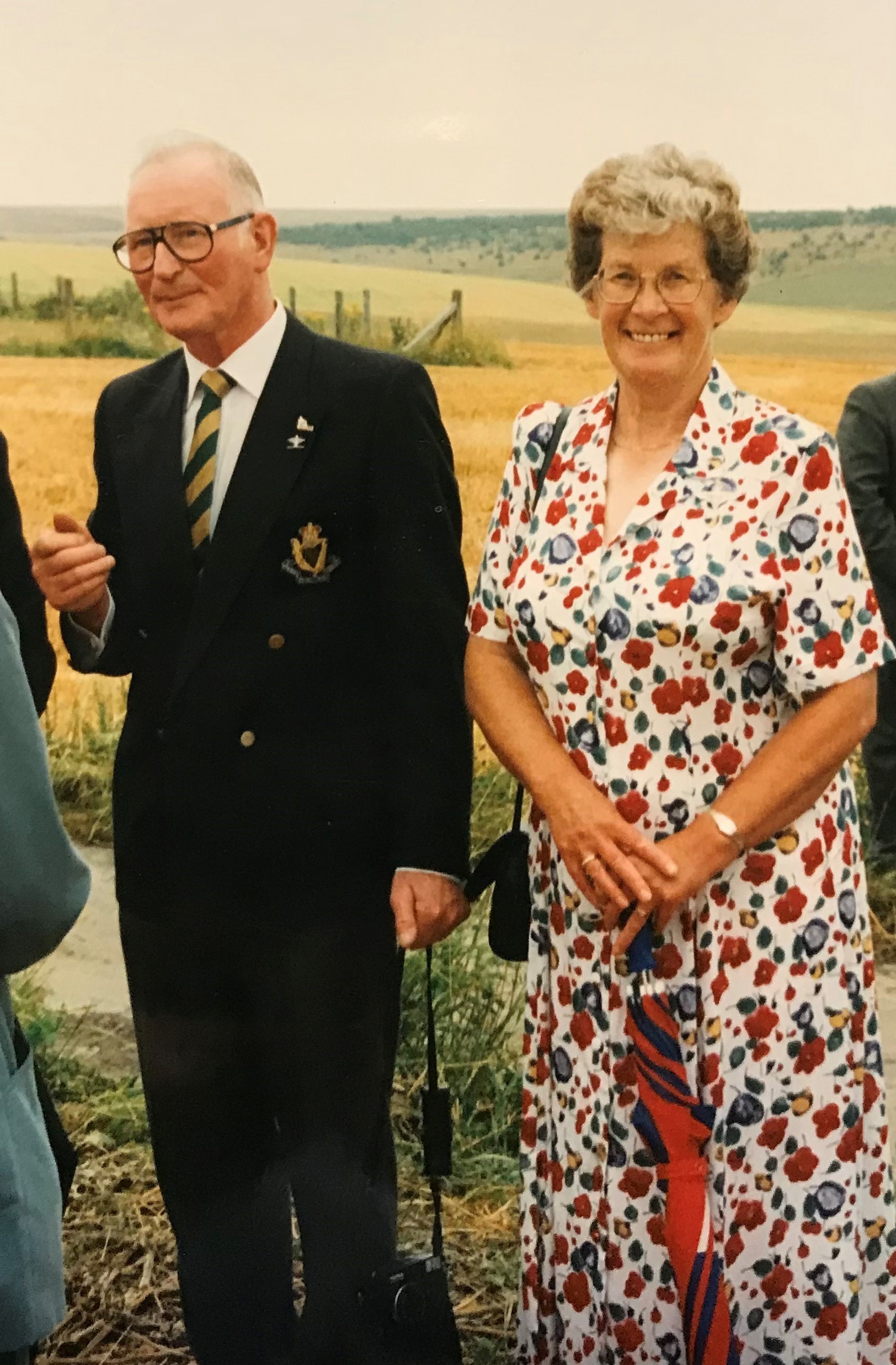 Elderly couple standing together outdoors, the man wearing a dark suit and glasses, and the woman in a floral dress, smiling in a rural setting with fields in the background.