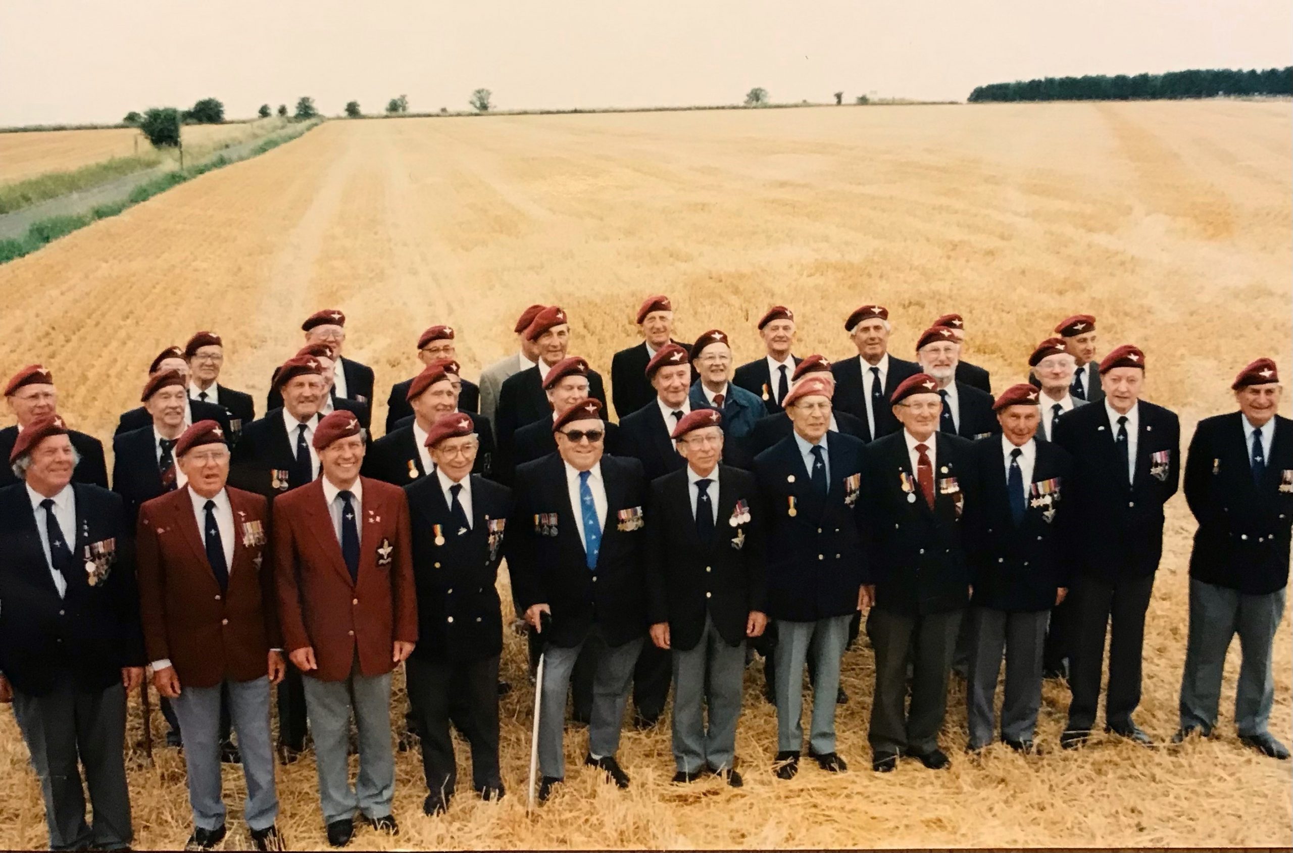 Group of men in formal attire standing in a wheat field, showcasing camaraderie and teamwork, with a backdrop of rural landscape.