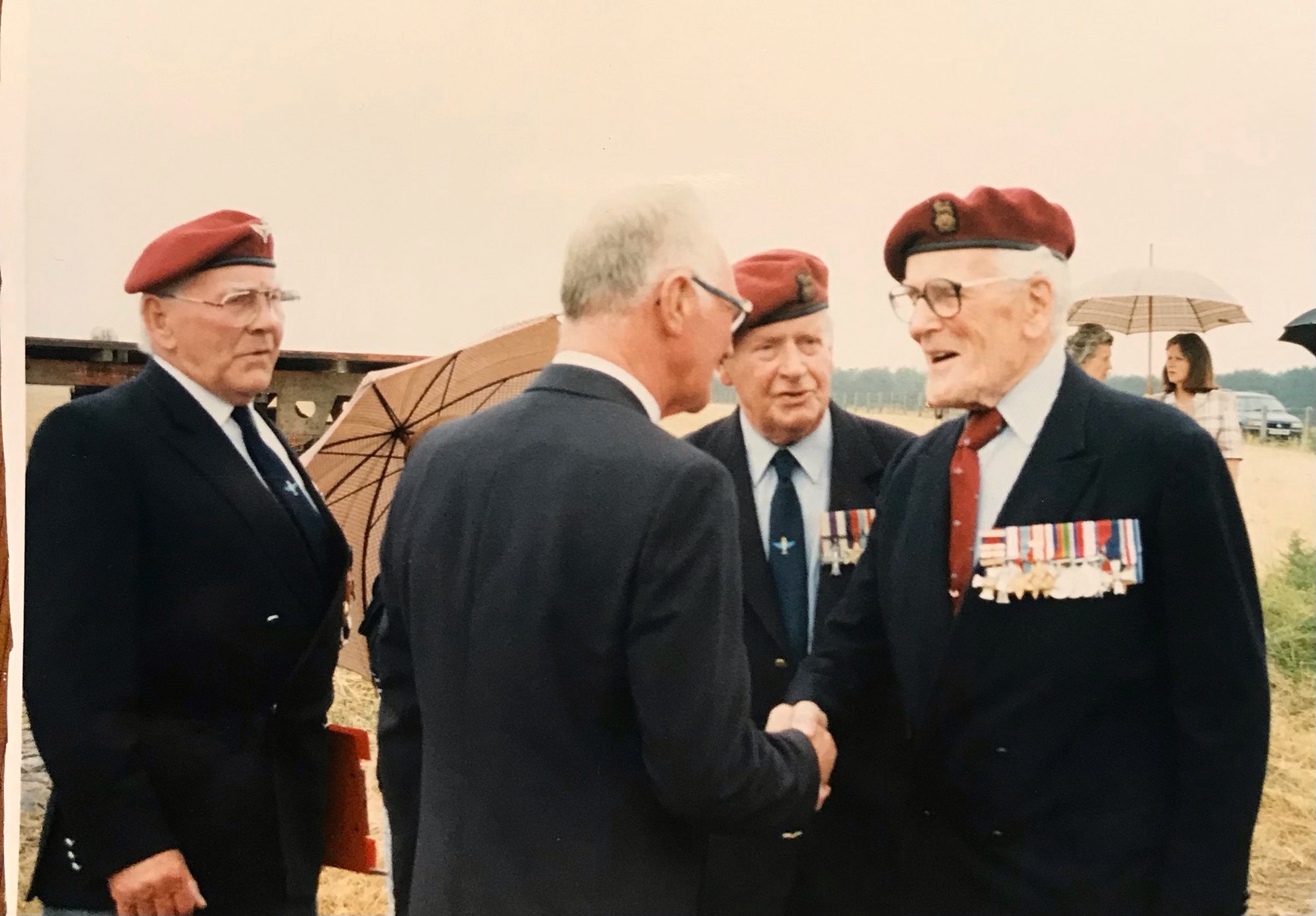 A group of four men, all wearing military uniforms and berets, engaged in conversation outdoors. One man is shaking hands with another, while the others observe. Several medals are visible on the uniforms.