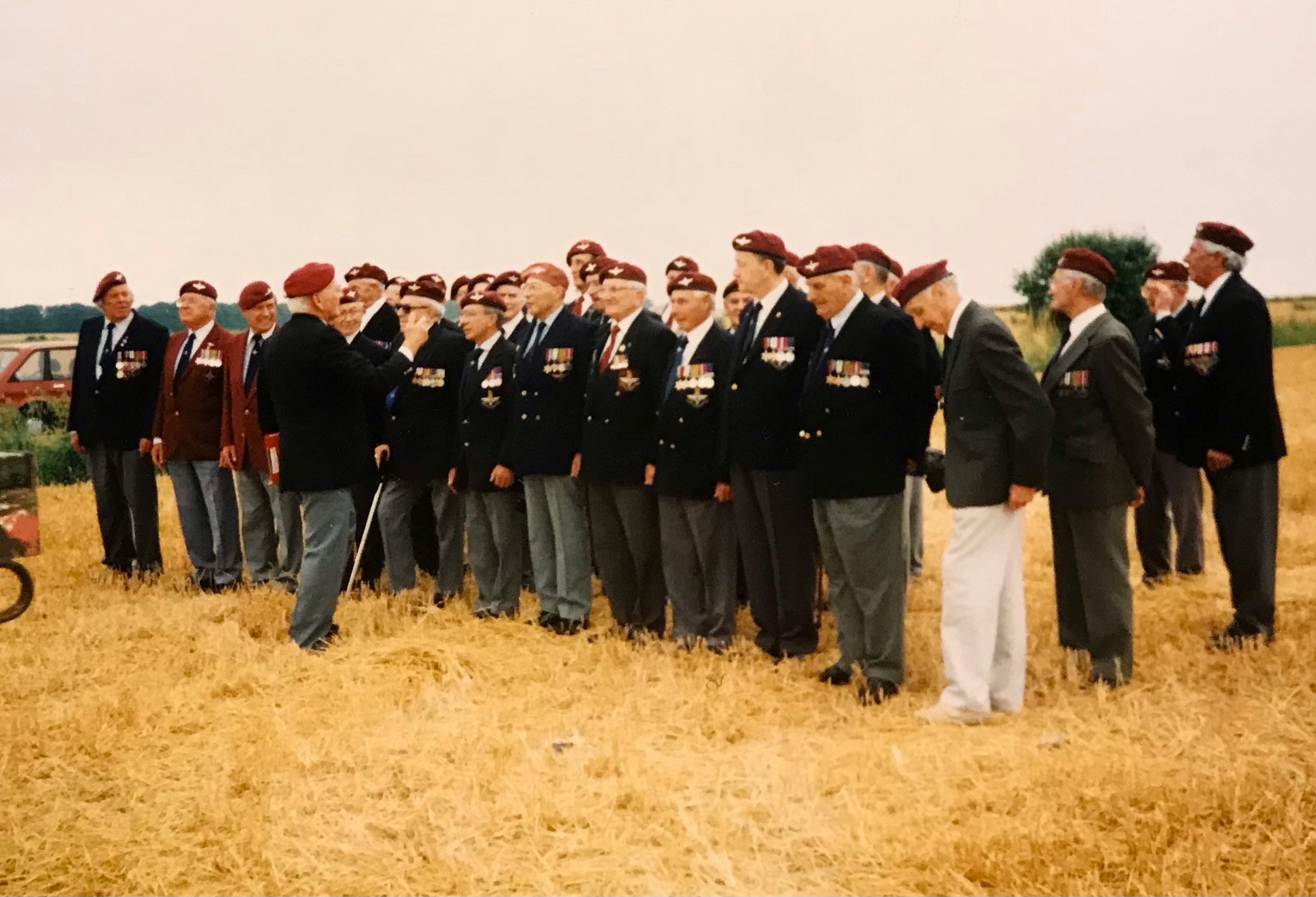 Group of veterans in military uniforms and berets standing in a field during a commemorative event, showcasing camaraderie and respect for their service.
