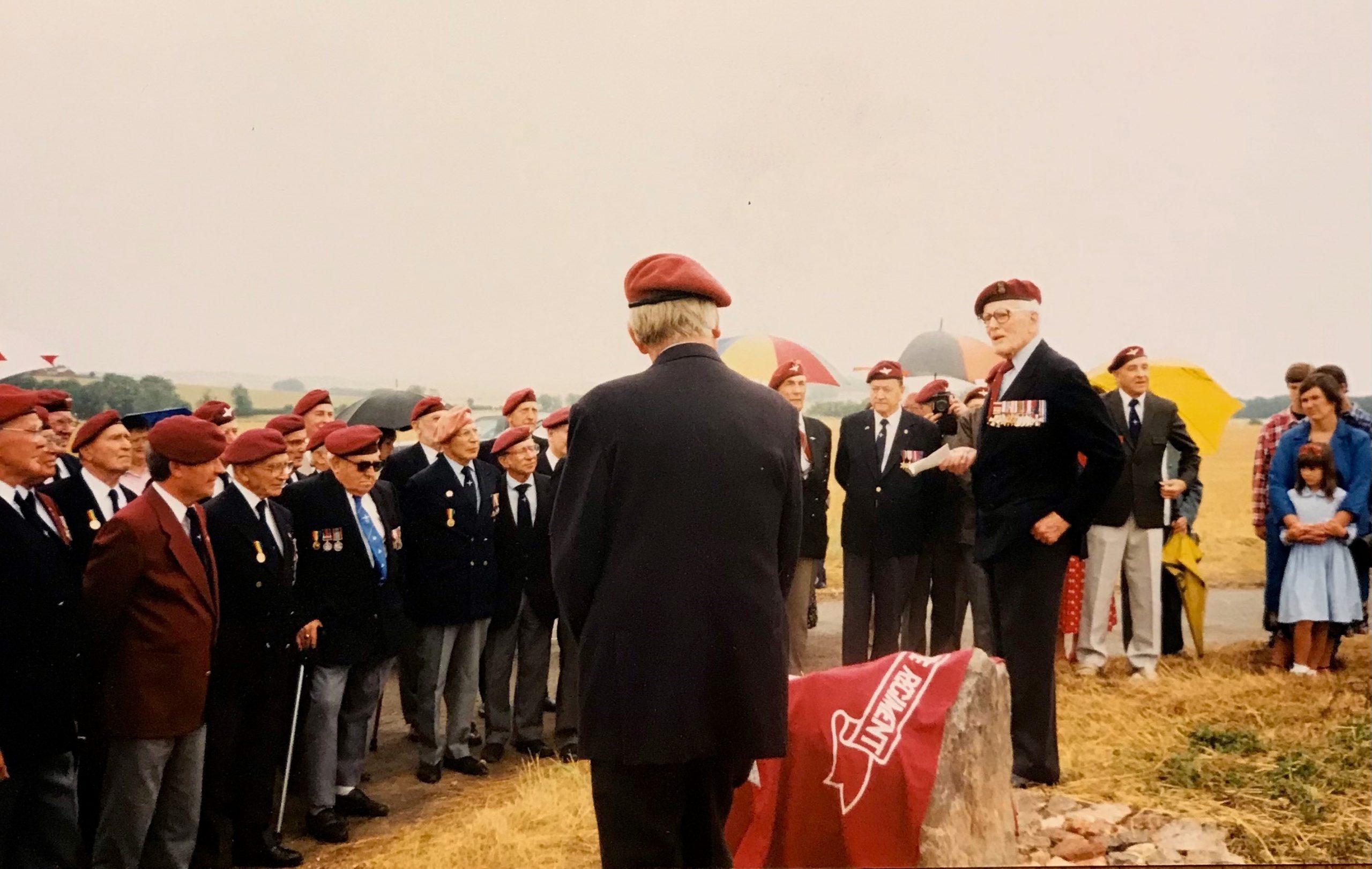 A gathering of individuals wearing red berets, attentively listening to a speaker at an outdoor event, with a backdrop of grassy fields and a cloudy sky.