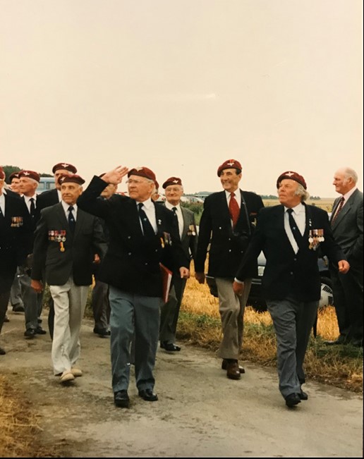 Group of veterans in formal attire walking together, some saluting, during a commemorative event outdoors.
