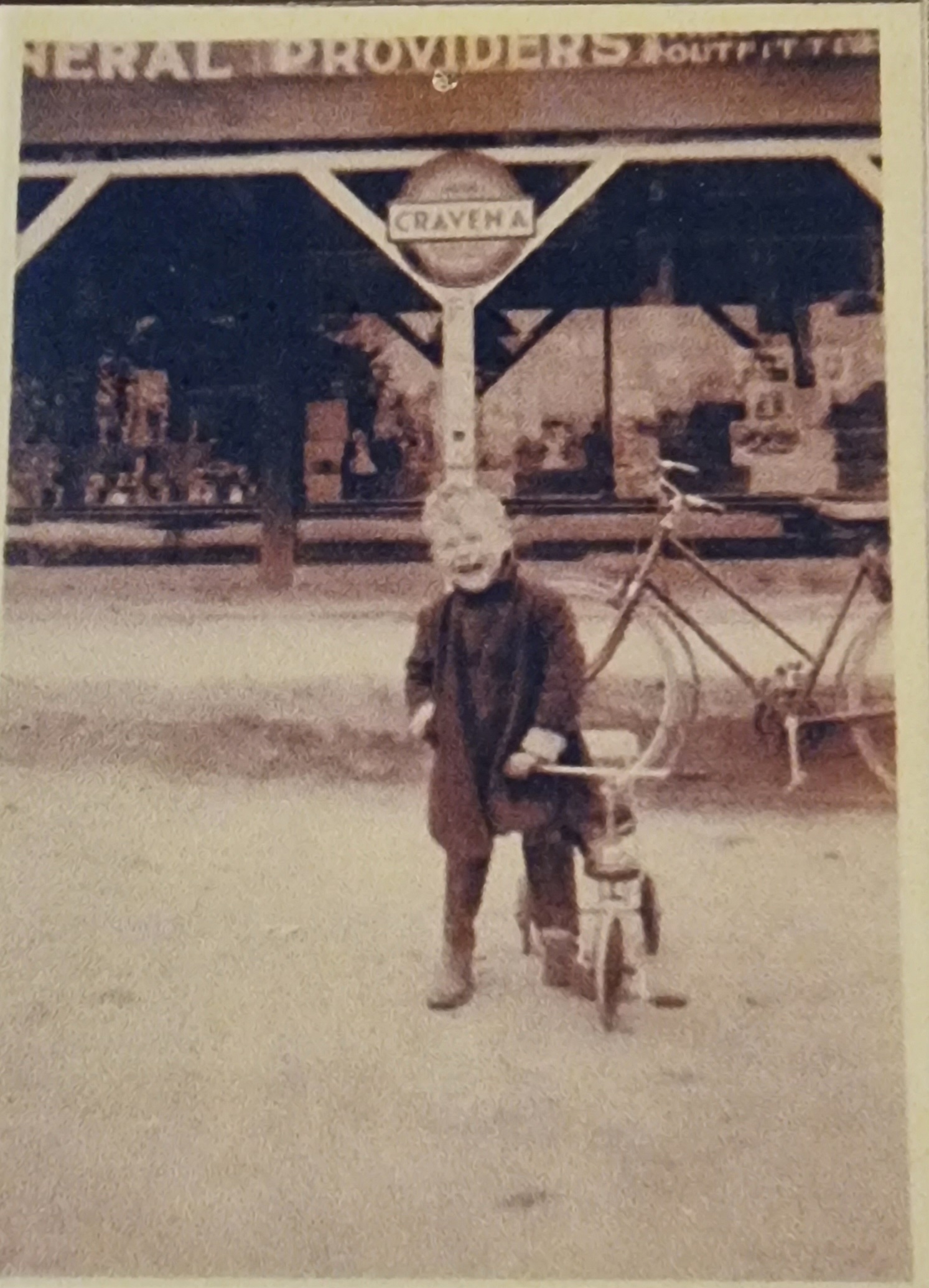 Historical photograph of a child standing beside a tricycle in front of a vintage general store, showcasing early 20th-century Americana. The store’s signage is partially visible, adding context to the time period.