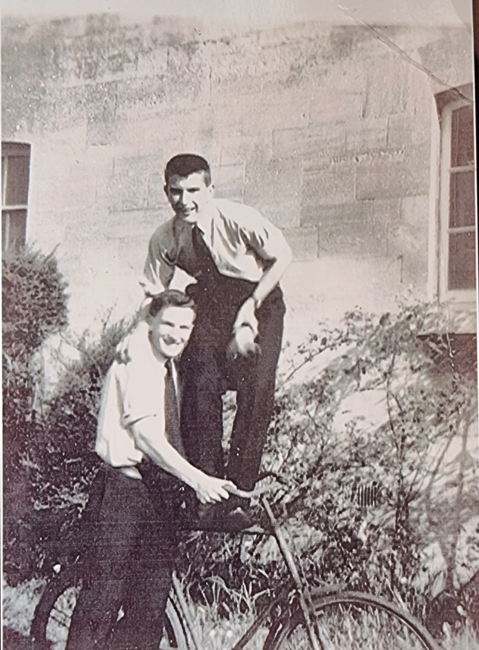 Two men posing with a bicycle in a vintage black and white photograph, one sitting on the bike and the other standing on the handlebars, surrounded by greenery and a building in the background.