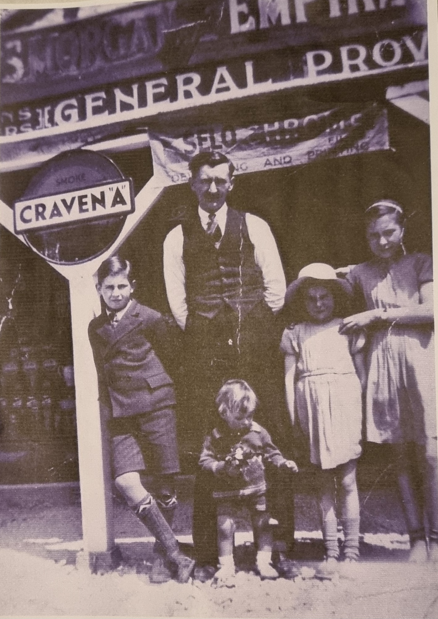 Historical black and white photo of a man in a vest and children, including a toddler, standing outside a general store marked "Craven." The backdrop features a vintage sign for "Empire General Provisions," reflecting early 20th-century retail.