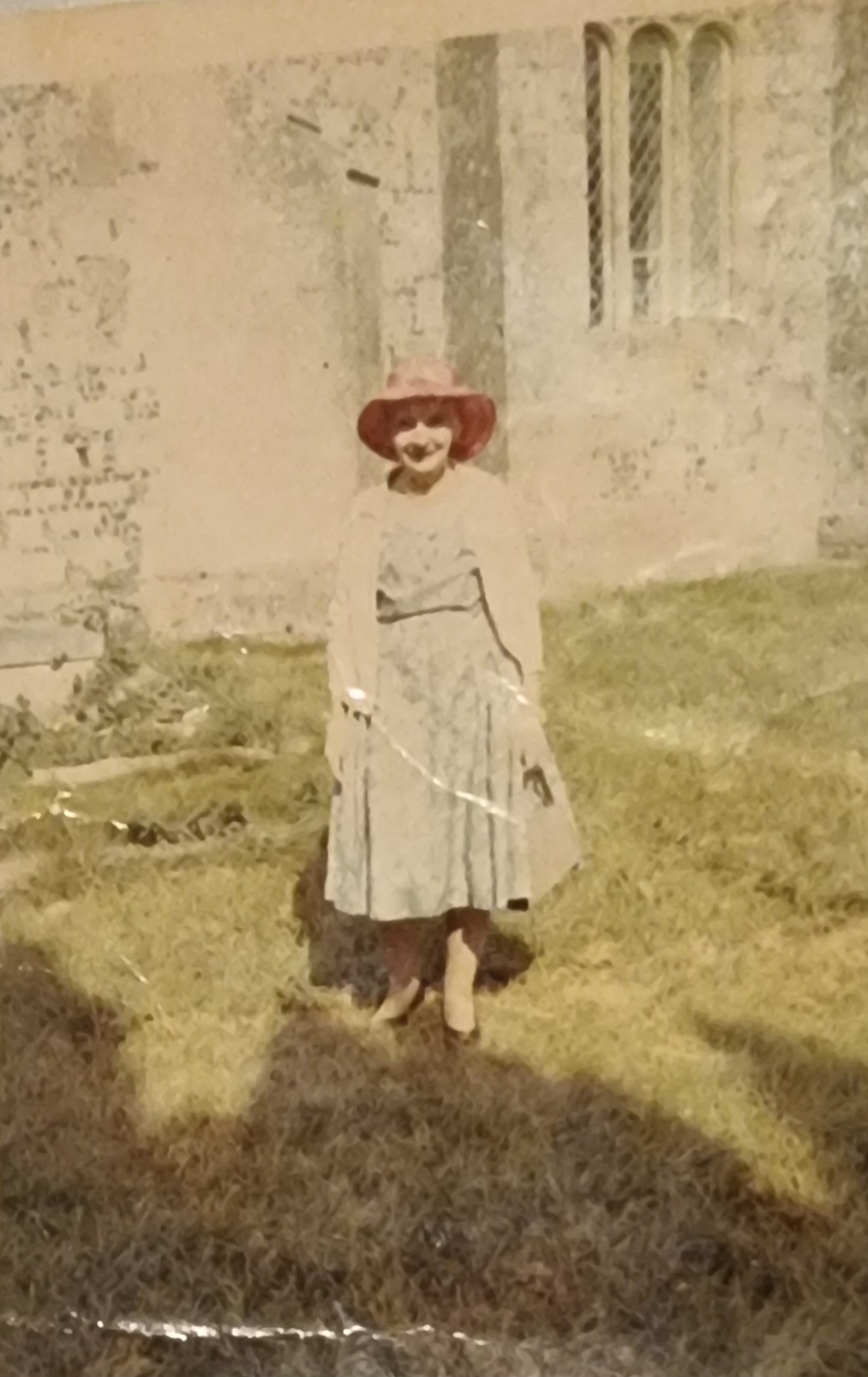 A young girl wearing a vintage dress and a wide-brimmed hat stands in a grassy area near an old stone building, showcasing early 20th-century fashion and architecture.