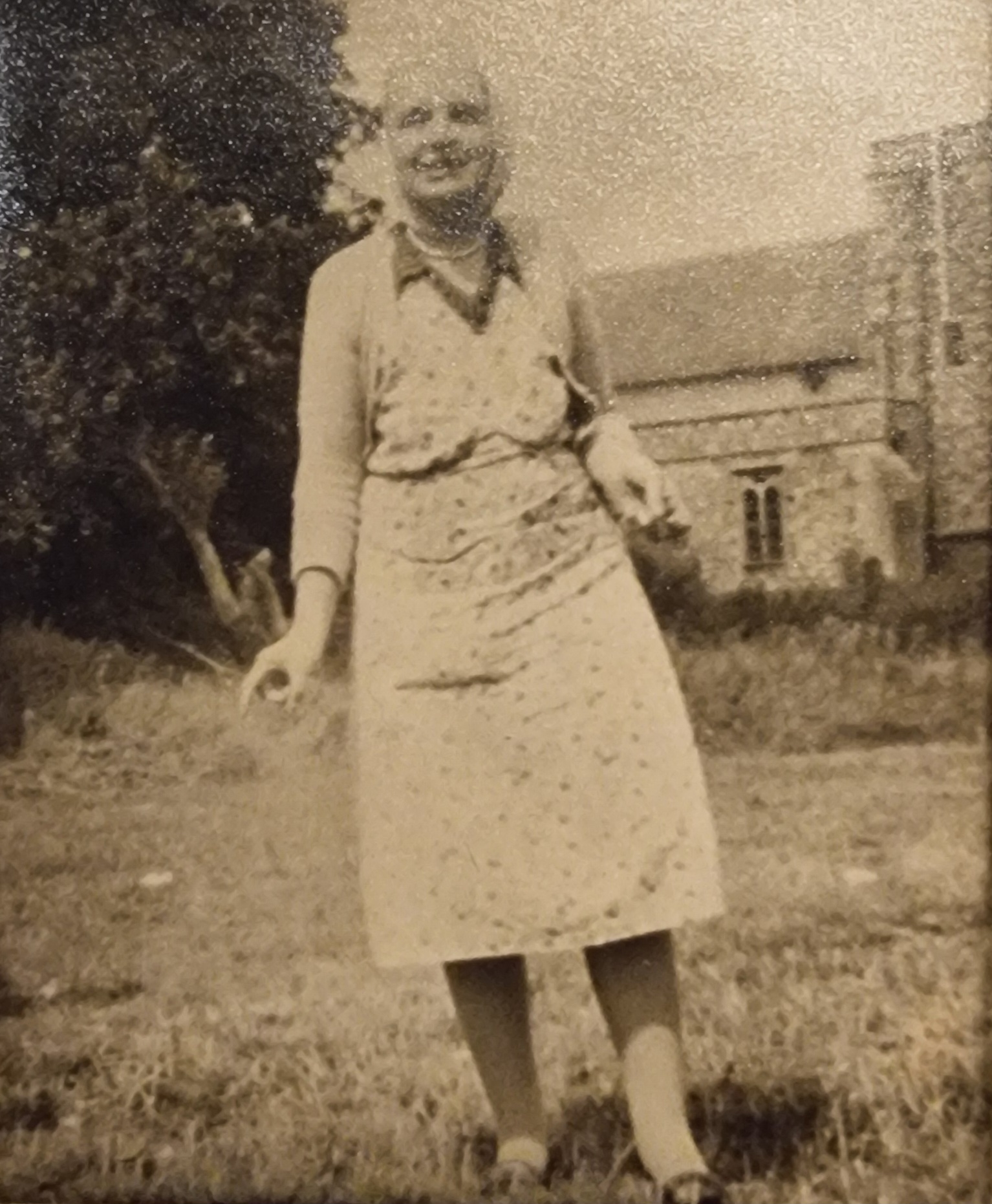 Elderly woman in a vintage dress standing in a grassy field, with a historic building in the background. The image has a sepia tone, suggesting an earlier time period.