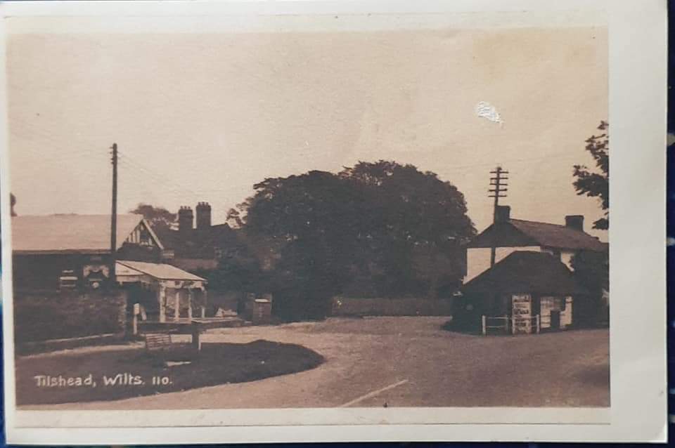 A vintage black and white photograph showing the intersection of two roads in Tilshad, Wiltshire. A small building with a sign and trees can be seen on the right, with more buildings in the background.