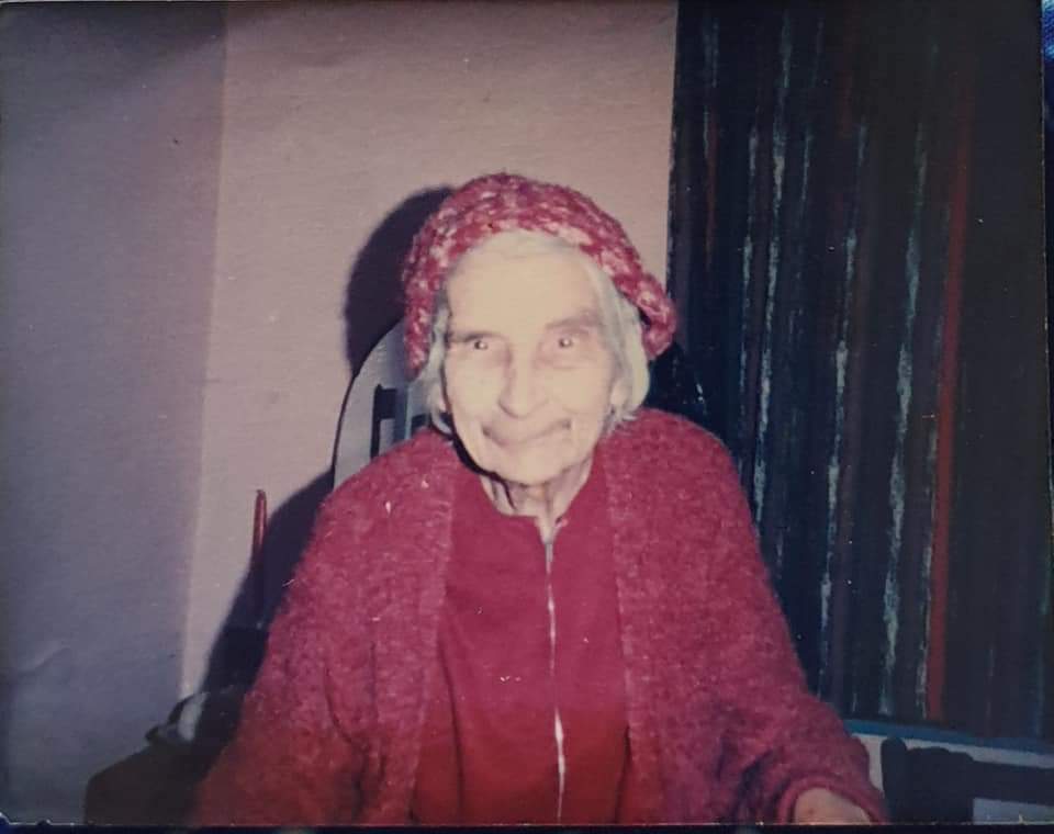 An elderly woman with gray hair wearing a red knitted hat and a matching sweater, sitting at a table with a warm smile. The background features a softly lit room with curtains.