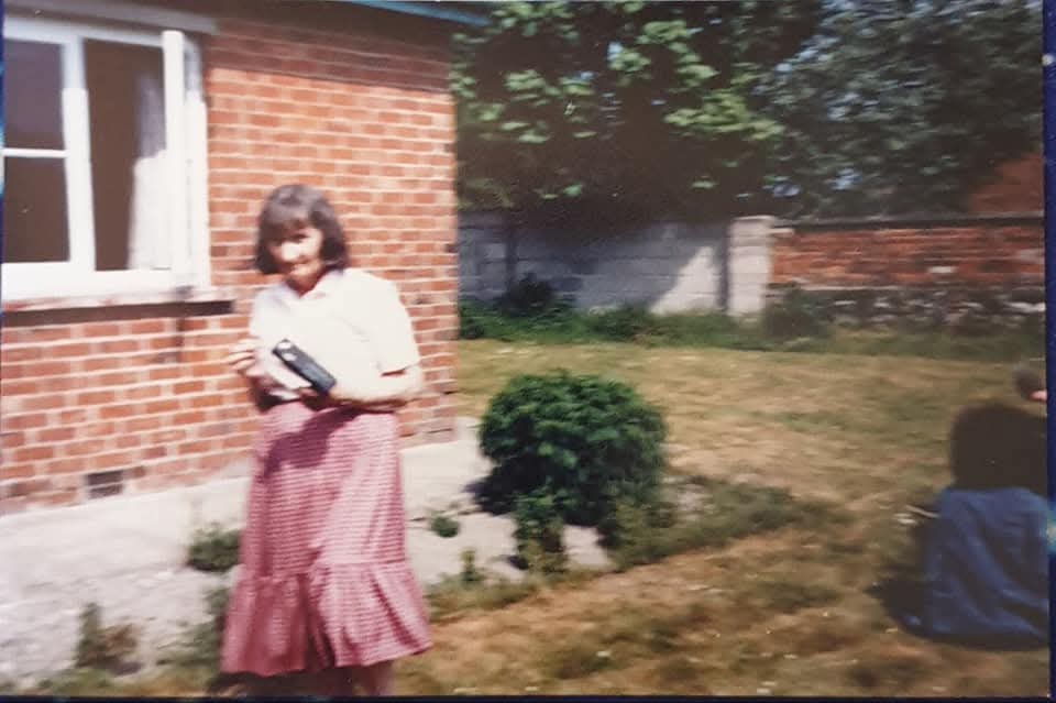 A young girl in a pink skirt stands in a grassy yard next to a brick house, holding a book, while a child sits in the background. The scene features trees and a sunny day, evoking a nostalgic atmosphere.