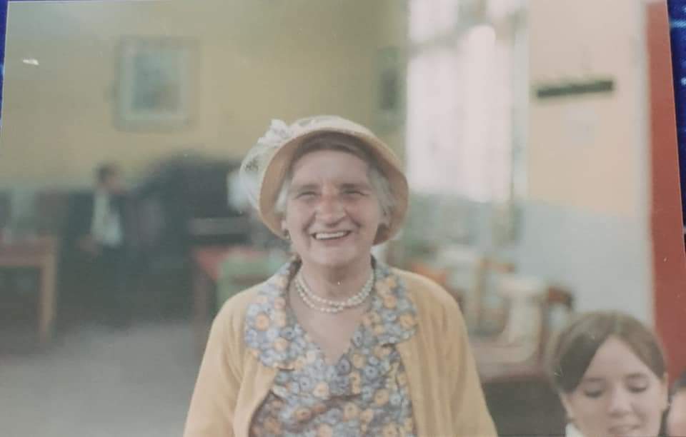 Happy elderly woman wearing a hat and yellow cardigan, smiling in a bright, cozy indoor setting with a young girl beside her.