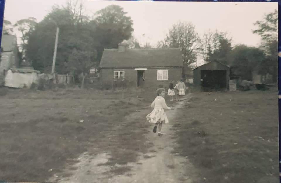 Children playing in a rural setting, running along a dirt path towards a small house and shed, surrounded by trees and open grassland. The image is in black and white, evoking a nostalgic atmosphere of a past era.