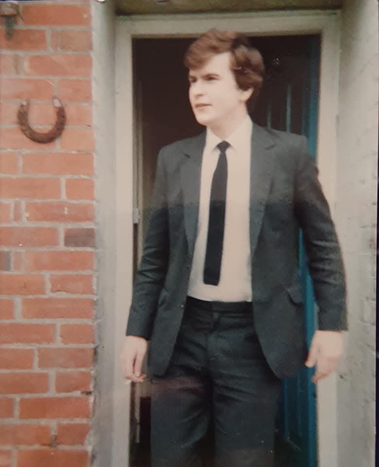 A young man in a dark suit and tie standing in a doorway, with a brick wall behind him. He has short, wavy hair and is looking to the side.