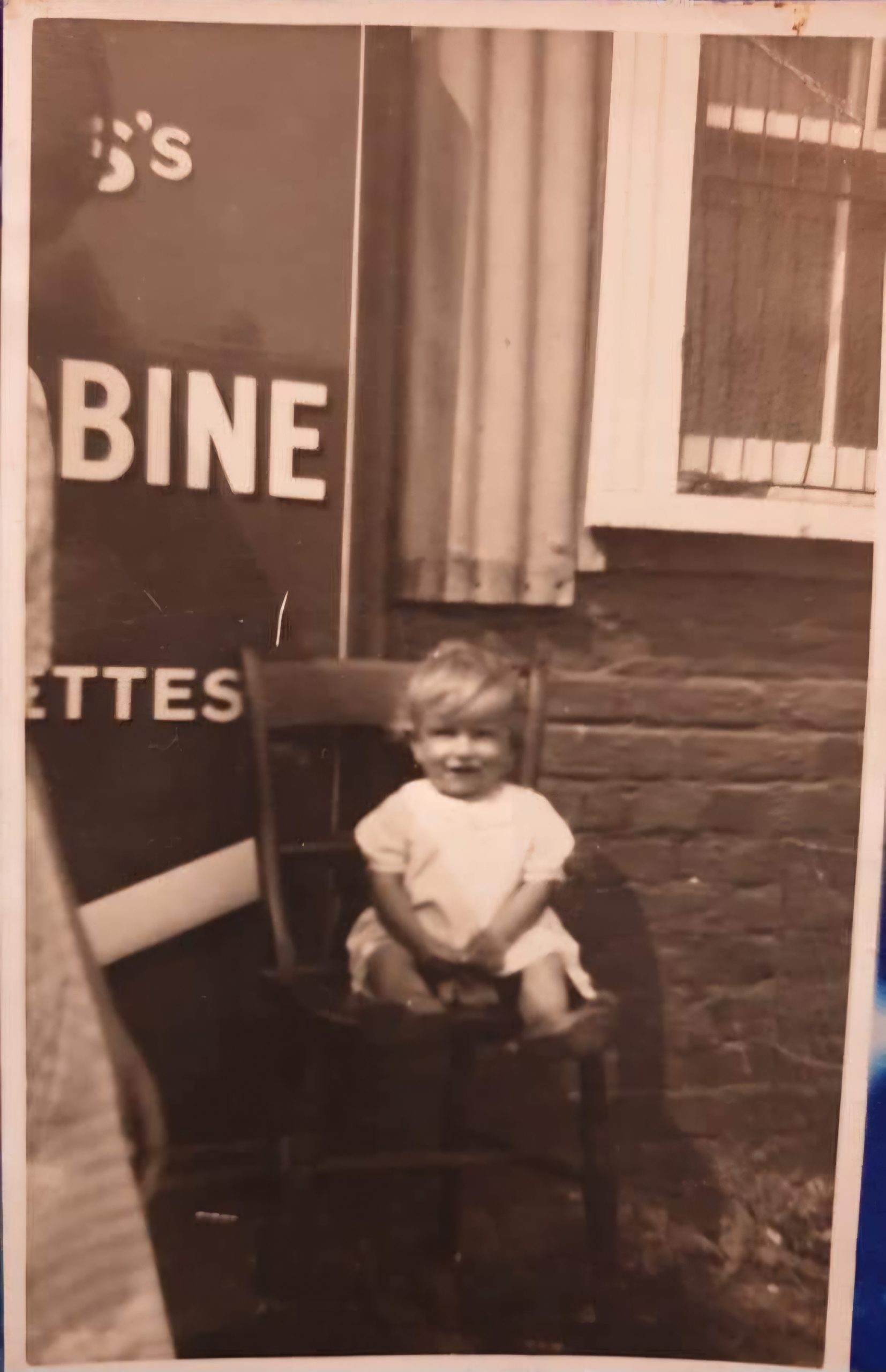 A black and white photograph of a small child sitting on a wooden chair in front of a brick wall with a sign that partially reads "Cigarettes." The child is smiling and wearing a light-colored outfit.